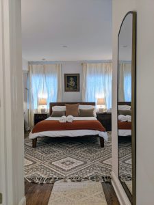 Centered view of the queen bed in Room 16 at Stanton House Inn, with antique portrait above the headboard, linen curtains, and wool area rug