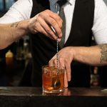 Barman in a black vest mixing craft cocktails in a speakeasy lounge near me