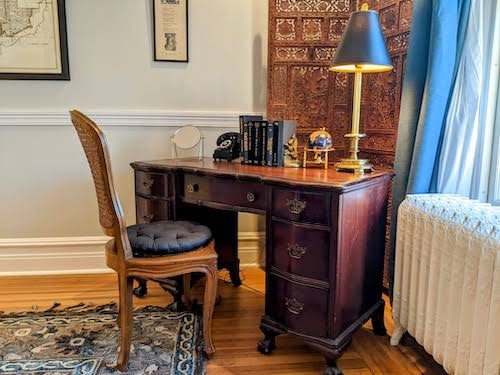A writing desk in a lived‑in guest room with worn wood floors: lamp on, chair pulled out